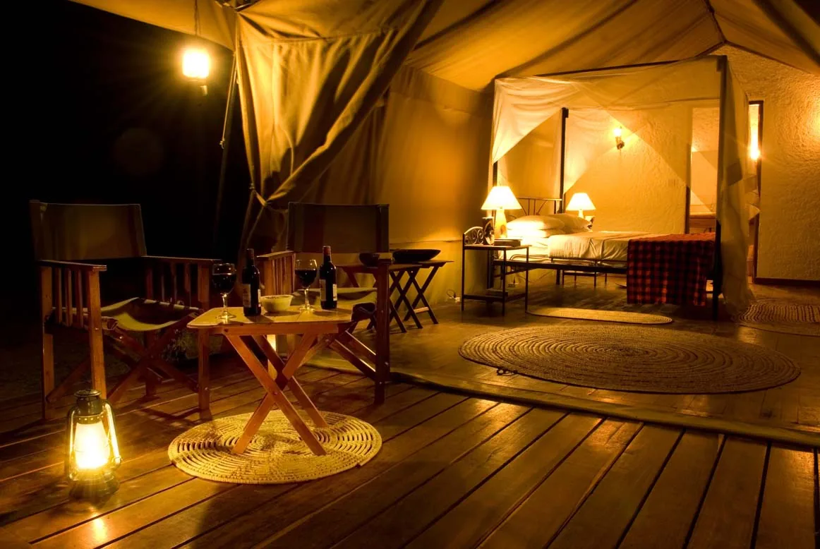 Inside a luxury safari tent at night, with lamplit bed, hardwood deck and director’s chairs