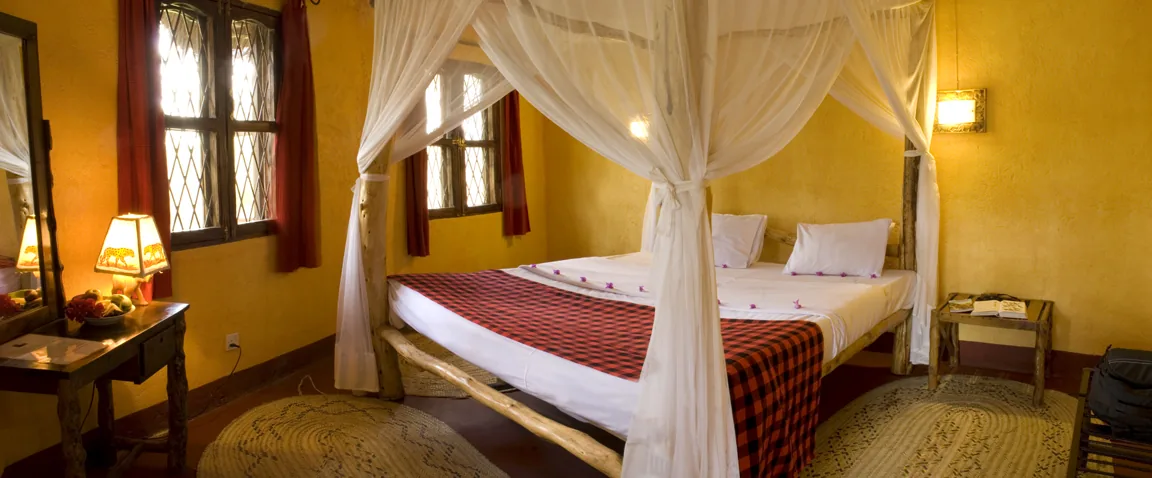 A double cottage room with a four-poster bed under mosquito netting, ochre walls and carved windows